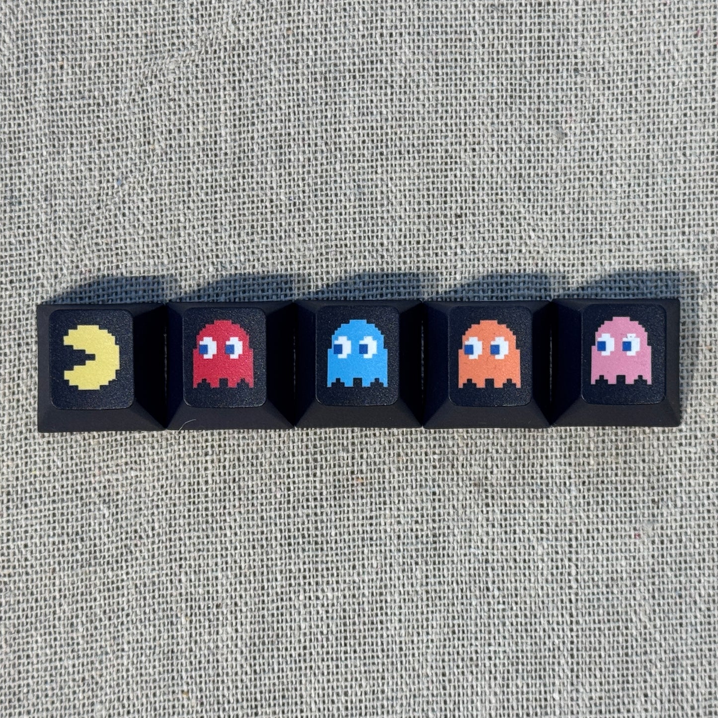 Five black Pac-Man keycaps featuring Pac-Man and the four ghosts Blinky, Inky, Clyde and Pinky arranged in a row.