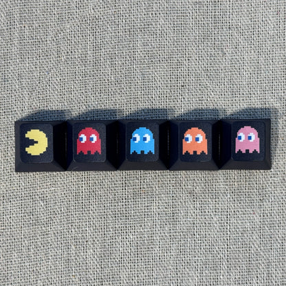 Five black Pac-Man keycaps featuring Pac-Man and the four ghosts Blinky, Inky, Clyde and Pinky arranged in a row.