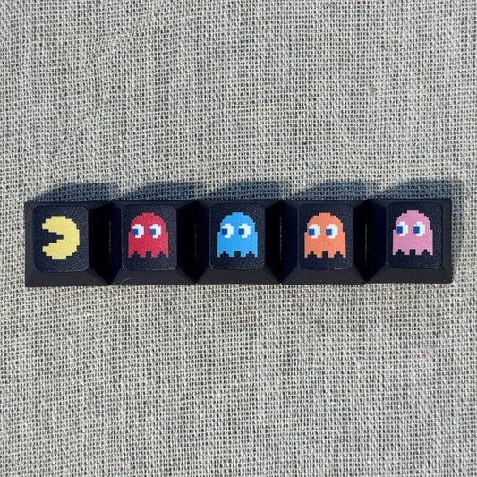 Five black Pac-Man keycaps featuring Pac-Man and the four ghosts Blinky, Inky, Clyde and Pinky arranged in a row.